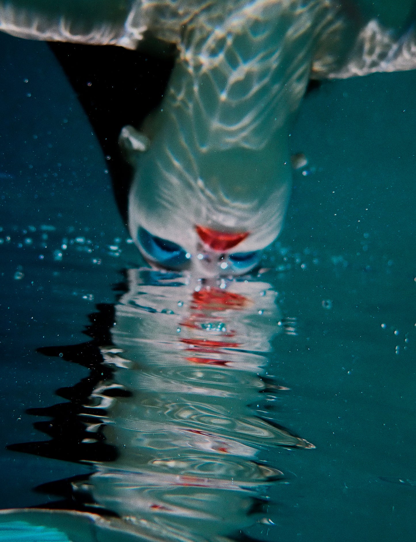 An underwater fine art photo print depicting a person with blue eyeshadow, partially submerged in water with air bubbles and a reflection on the surface.
