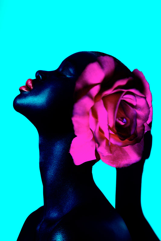 Profile portrait of a black bald woman with natural hair and pink lipstick with a pink rose collage on her head set against a vibrant teal background.