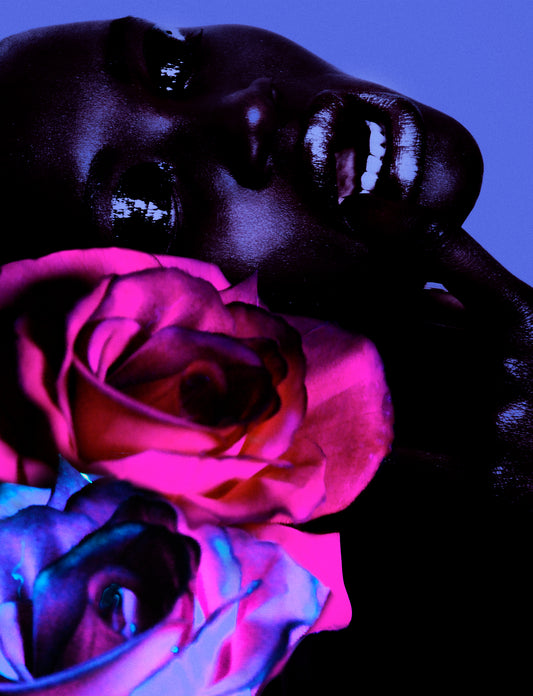A collage of neon pink and purple blue roses are in the foreground. Behind them is a portrait of a dark black model with her head tilted back and mouth open. She is wearing glossy black eyeshadow on her eyelids.