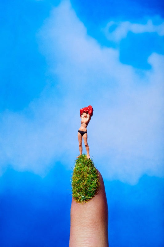 An artistic photograph featuring a miniature figure of a topless woman in a playful pose as she flashes takes her shirt off atop a grassy fingernail set against a beautiful blue sky with clouds.