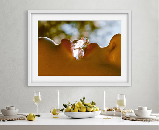 Framed artwork of a person holding an ice cube in their mouth, displayed above a dining table with fruit and glasses.