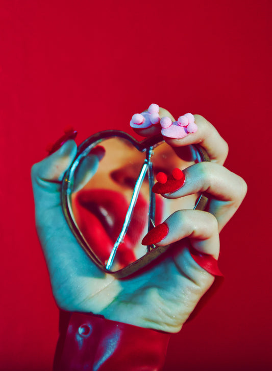 valentine's day love image of woman's hand with pom-pom nails manicure holding broken shattered heart mirror with lip reflection red background