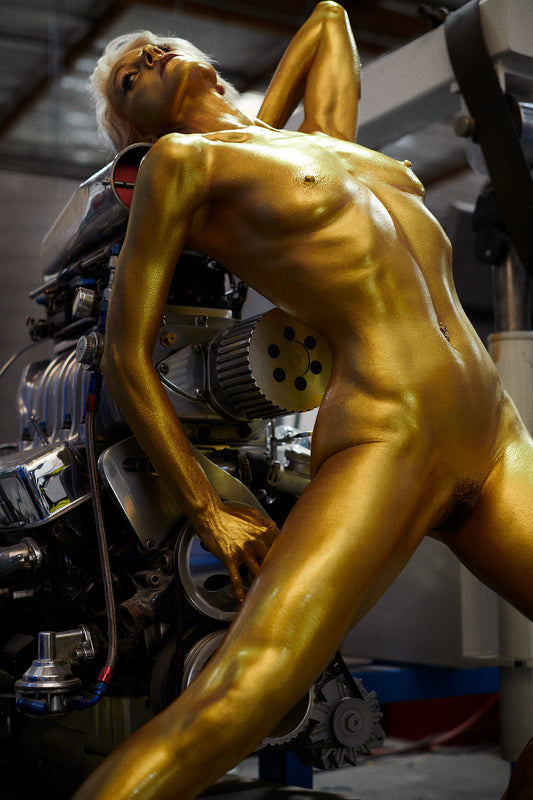 Gold-colored sculpture of a nude figure leaning against a large supercharged engine.