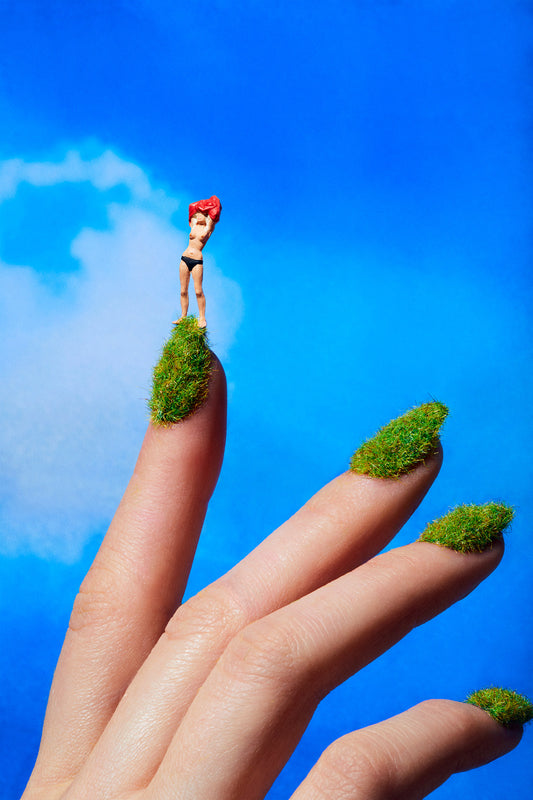 Miniature dollhouse figurine woman on top of a grassy hill that is actually a woman's fingernails covered in grass nail polish. The woman is flashing the camera taking her shirt off.