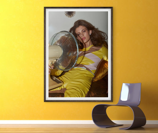 Framed artwork of a woman leaning against a fan on a yellow wall with a modern chair.
