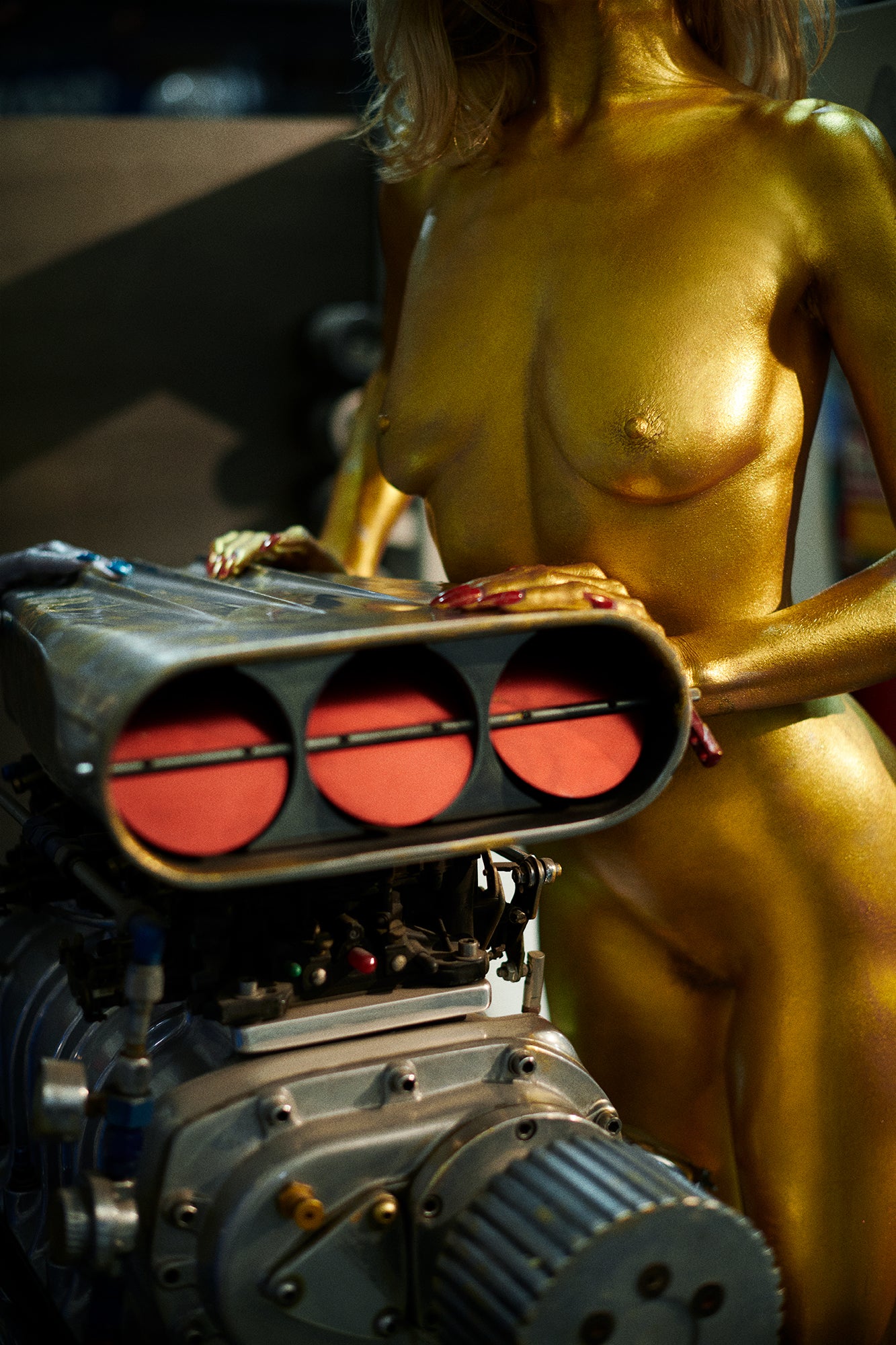 Nude woman painted gold looking like a Goldfinger bond girl standing near a supercharged mechanical car engine.