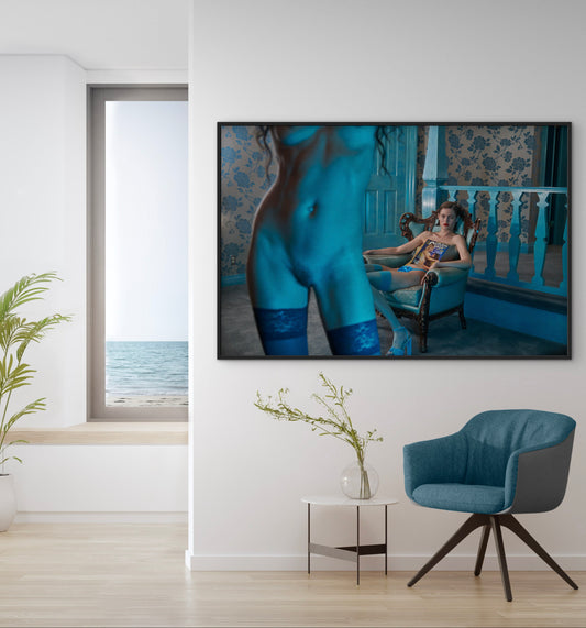 Framed artwork on a wall with a chair and table below in a room with a view of the ocean.