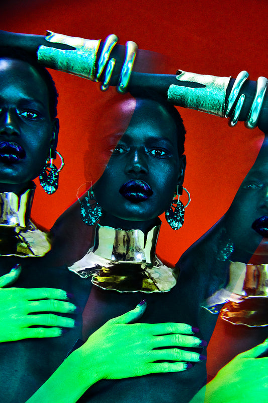 Black woman, model Ajak Deng is wearing gold jewelry with neon green body paint on her hand. The image is shot with a filter that has creative a multiple exposure effect, and there are three images of the woman's face