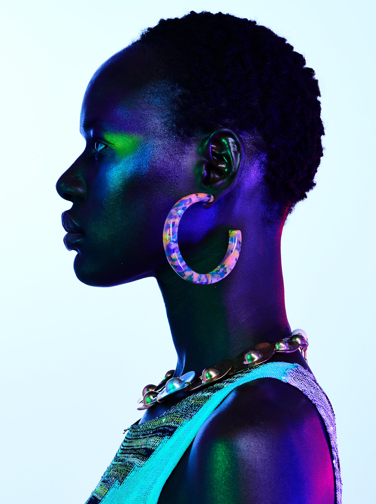 A profile portrait of a woman with a metallic eye shadow, blue fingernails, and large hoop earrings, against a colorful background.