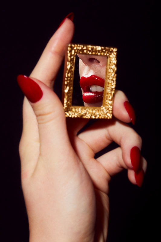 A person with red oval nails holding a small mirror with a gold frame, reflecting red lips.