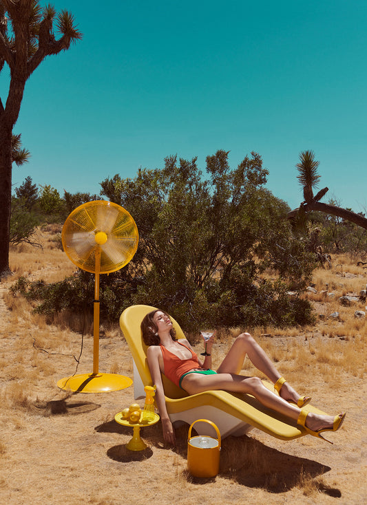 Woman lounging in a yellow chair with a vintage-style fan and drinks in a desert setting