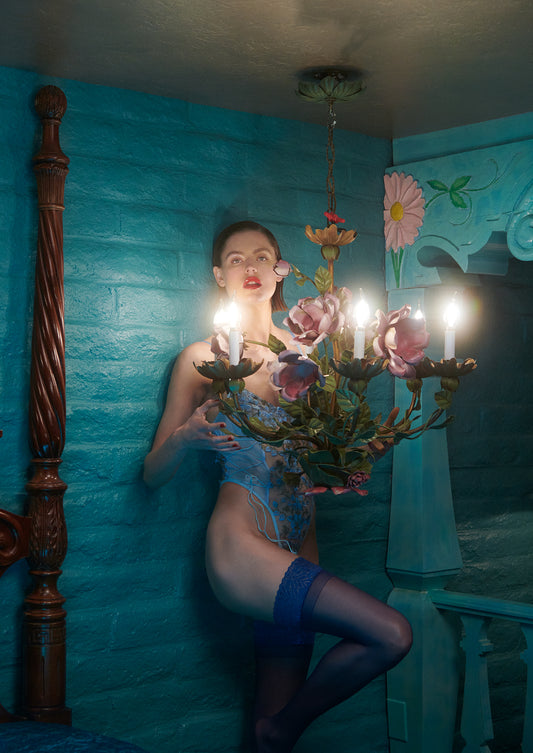 Woman in a blue bodysuit sitting under a chandelier with floral design against a teal wall.