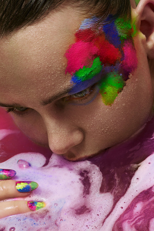 A close-up of a woman with colorful face paint and nails, partially submerged in a bathtub with pink foam and colorful water, featuring a vibrant, abstract multicolored pattern on her face.