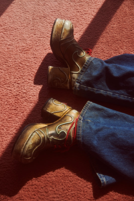 A pair of men's brown platform shoes with a heart pattern lying on a red carpet next to blue jeans, representing a retro fashion editorial theme.