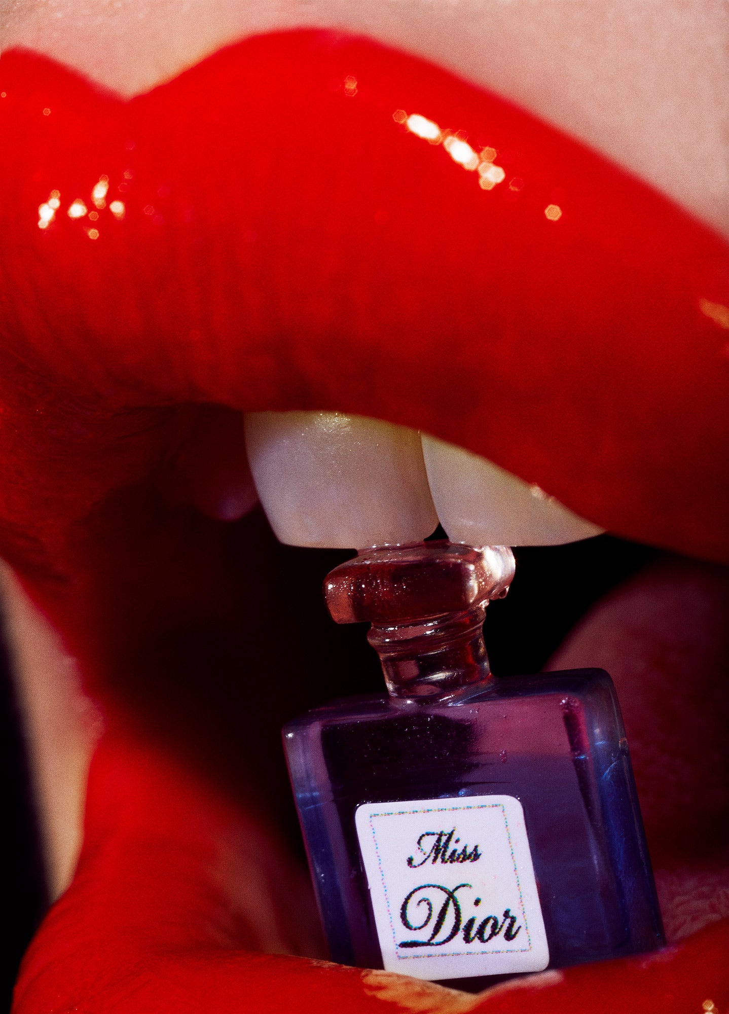 Close-up macro beauty portrait of red glossy plump lips with miniature Dior perfume bottle resting between teeth.