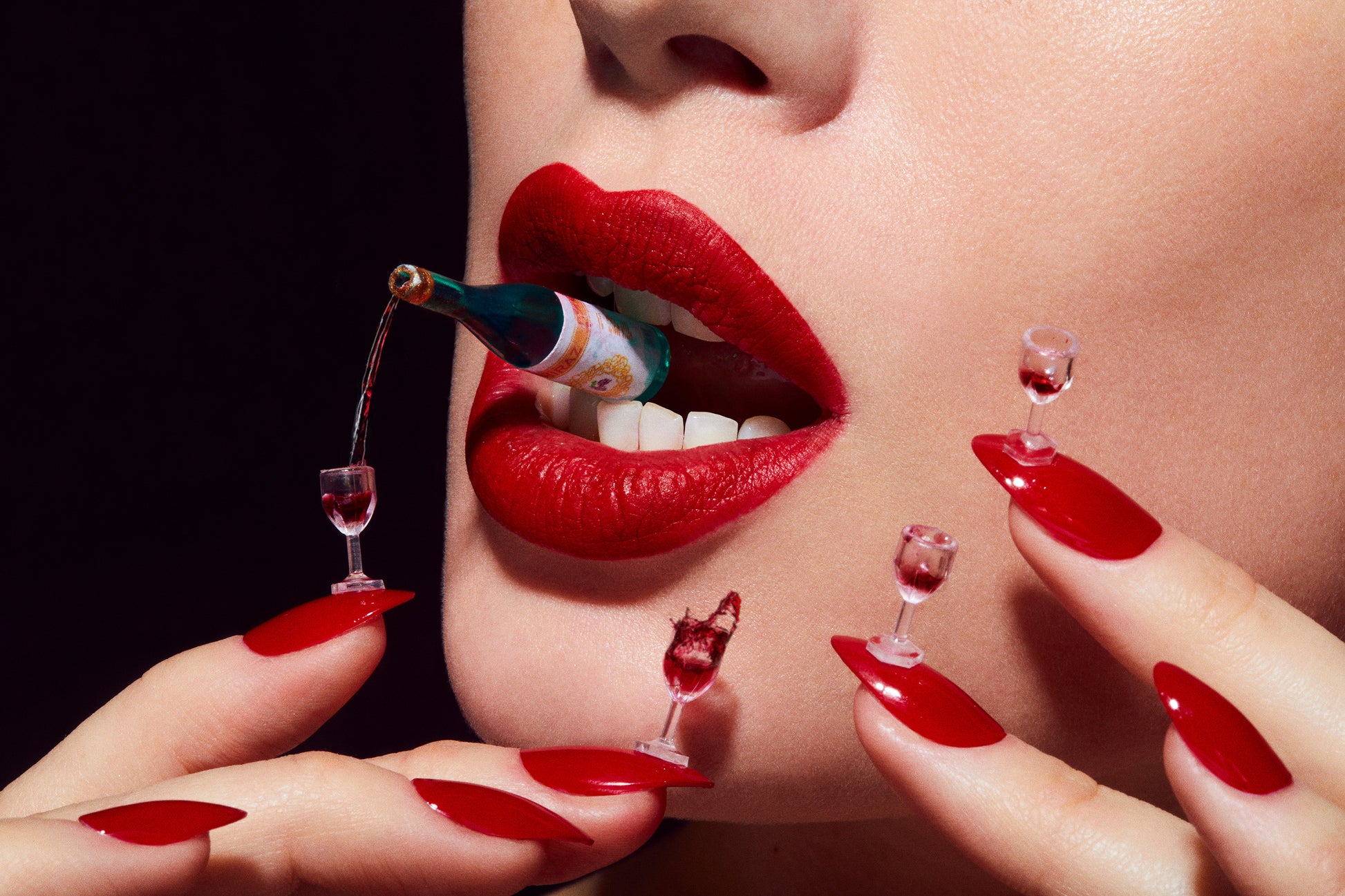 A close-up art photograph of a person with red lipstick and red fingernails, with miniature wine glasses on the nails. The image depicts a playful and celebratory theme.