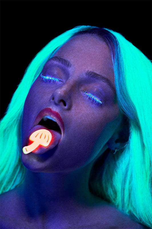 A person with glow-in-the-dark blue hair, wearing eyeliner, is depicted in a UV light with a glowing piece of candy in their mouth. The image has a neon aesthetic.