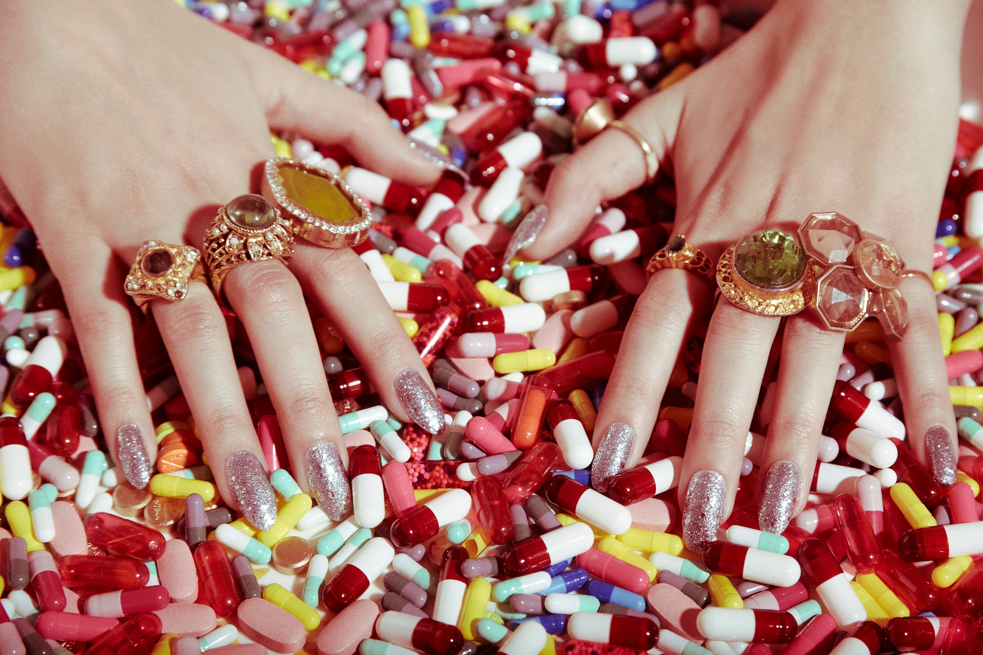 Vintage style close-up of a person's hands with multiple rings on fingers, surrounded by a colorful assortment of pills.