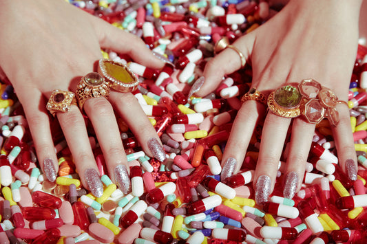 Vintage style close-up of a person's hands with multiple rings on fingers, surrounded by a colorful assortment of pills.