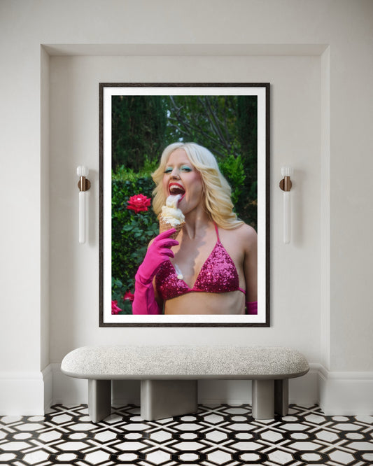 A fine art print of Amy Louise Taylor by Jamie Nelson. The punk singer of Amyl and the Sniffers has a blonde flip-out hairstyle and is wearing 80's style clothing and makeup—a pink bikini, gloves , blue eyeshadow, and red lipstick. She licks a messy ice cream cone with her big tongue as it drips onto her.