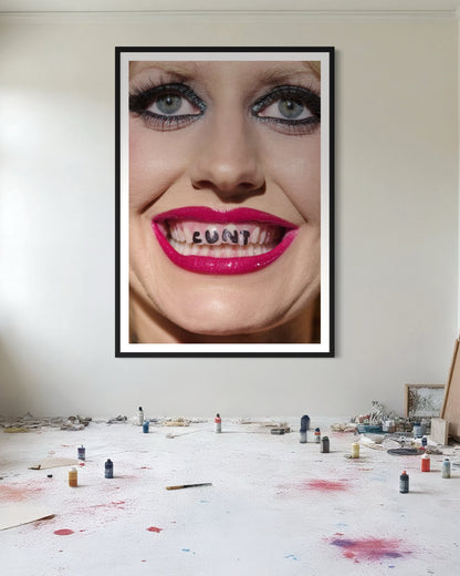 Framed artwork of a Amy Taylor's face shown smiling with 'CUNT' written on her teeth, on a wall above a table with art supplies in a messy painting room.