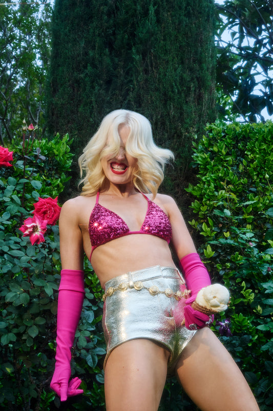 Amy Taylor (Amyl and the Sniffers) photographed by Jamie Nelson. She is sporting blond locks is styled in retro glam rock clothing including pink sequins and metallic hot pants. She is head banging and dancing as she grips an ice cream cone near her crotch giving "BDE".