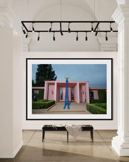 Framed photograph of a pink house with a punk style blonde woman (Amy Taylor of Amyl and the Sniffers band) flexing her muscles while wearing extra long jeans that go from the roof to the ground.