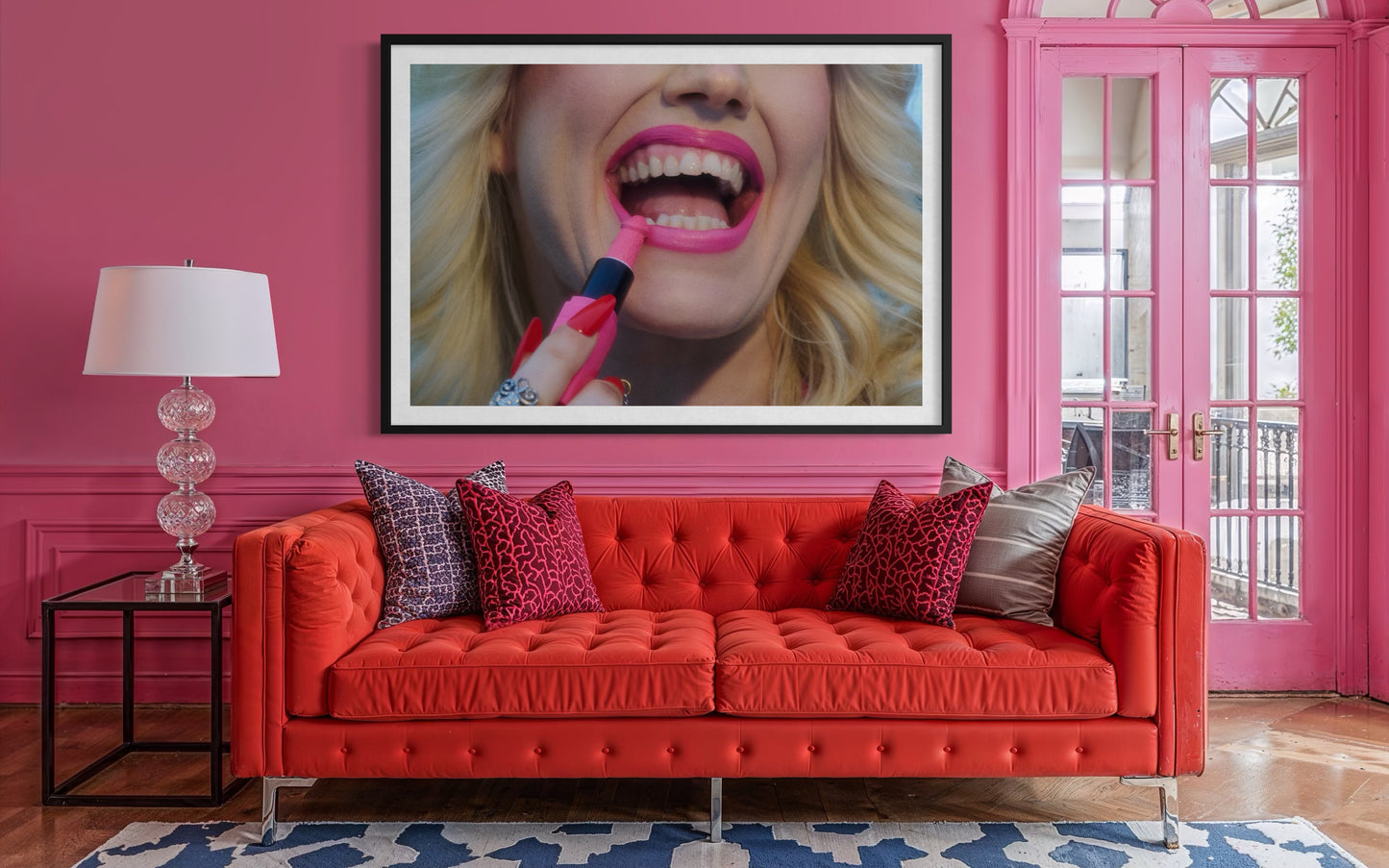 Amy Taylor by Jamie Nelson for Vogue Portugal. A large fine art print of a blonde woman smiling and laughing as she applies lipstick. The print is displayed in a pink Barbie styled interior living room above an orange couch.