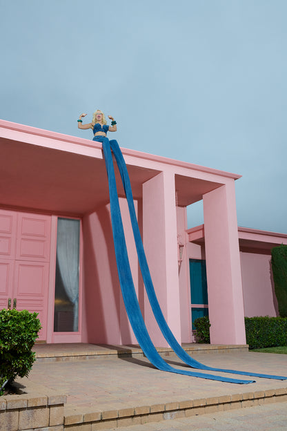 Blonde woman (Amy Taylor of Amyl and the Sniffers band) flexes her muscles while wearing the world's longest jeans on top of the roof of a pink mansion.
