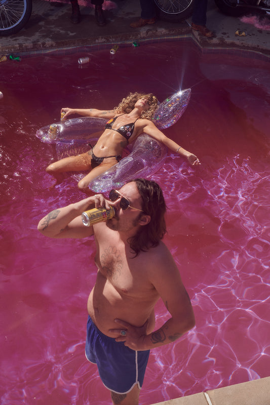 An art print depicting a whimsical scene with a woman lounging on a pink glittery pool float, inspired by 1970s and 1980s party culture, with a man drinking beer in the foreground. The pool water is pink, and the float appears to have glitter, adding a playful, psychedelic vibe to the image.