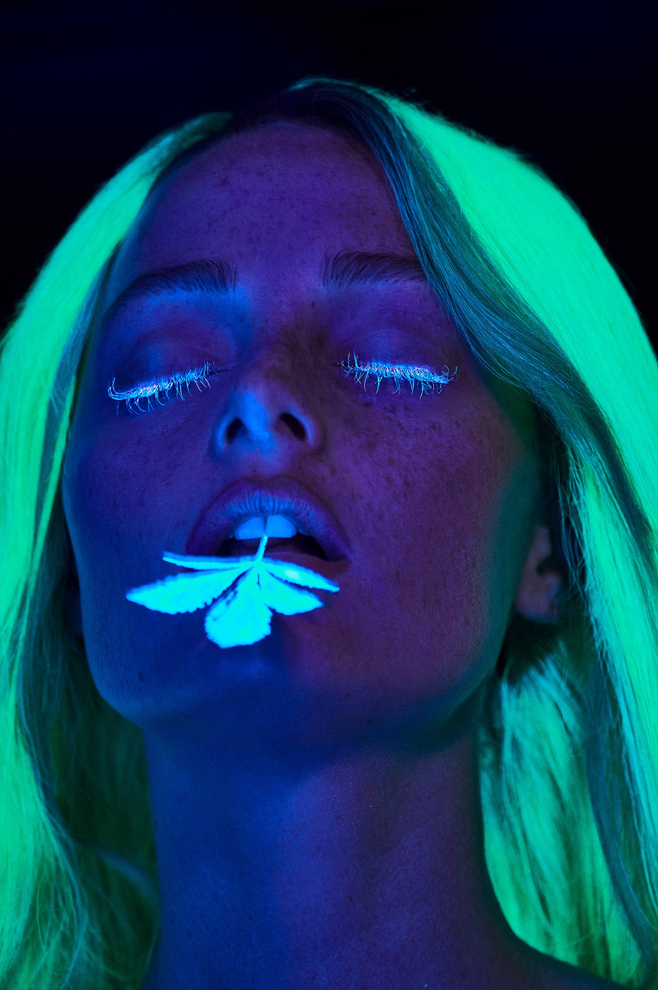 Artistic photography of woman with freckles in black light with neon green hair and white glowing eyelashes with marijuana pot leaf in her mouth