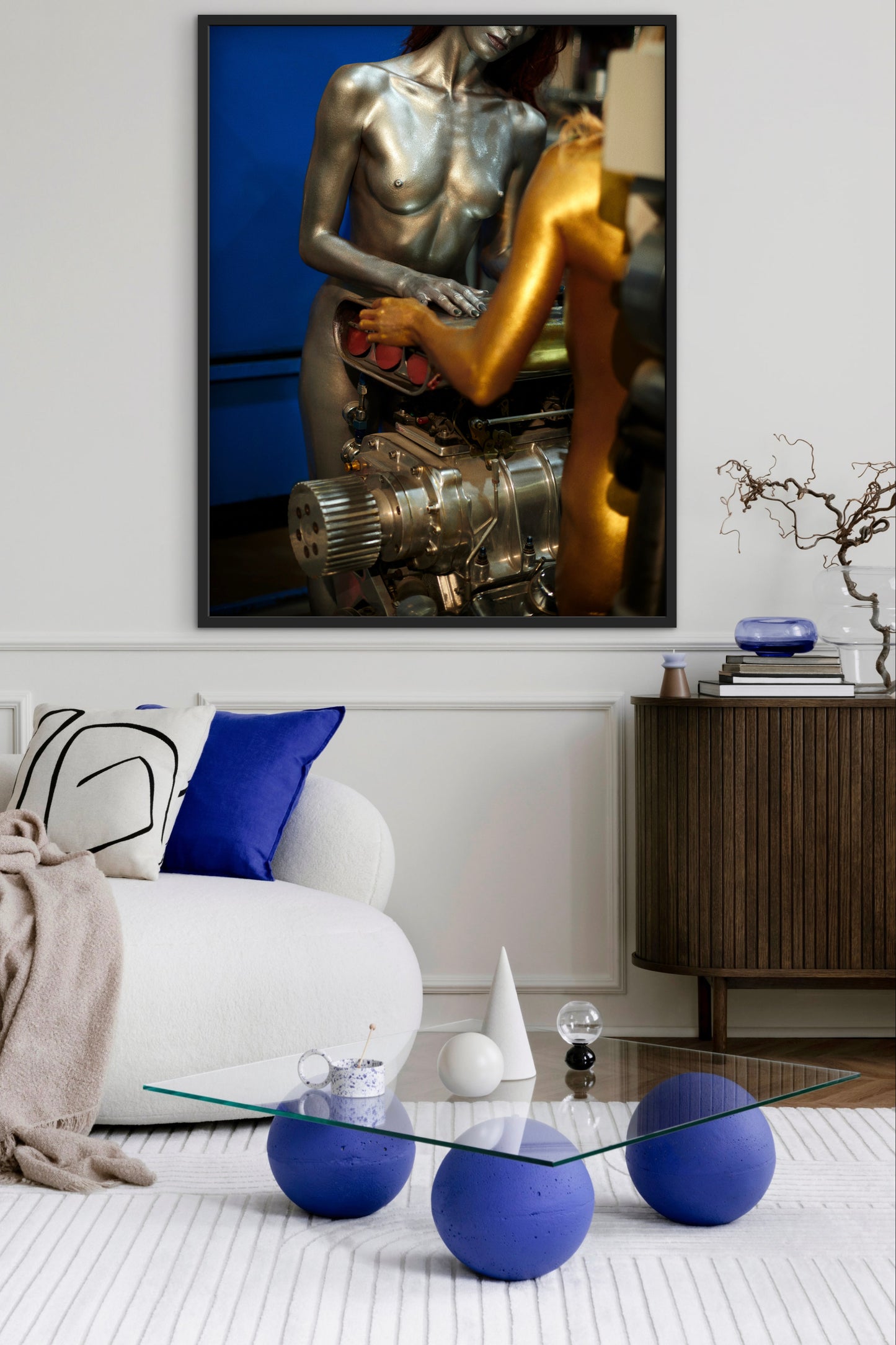 A modern living room with abstract art of two nude female mechanics working on a big block car engine. The room also has blue spherical ottomans and a glass coffee table.