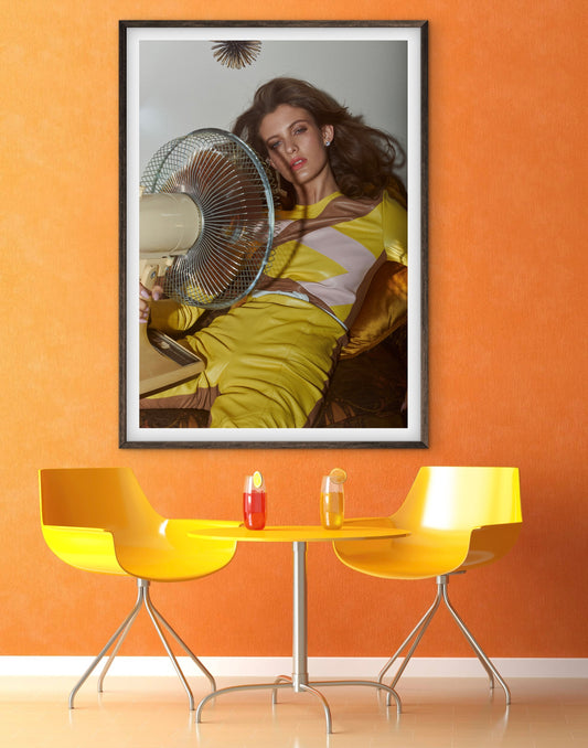 Framed artwork of a woman with a fan on an orange wall above a yellow table and chairs.