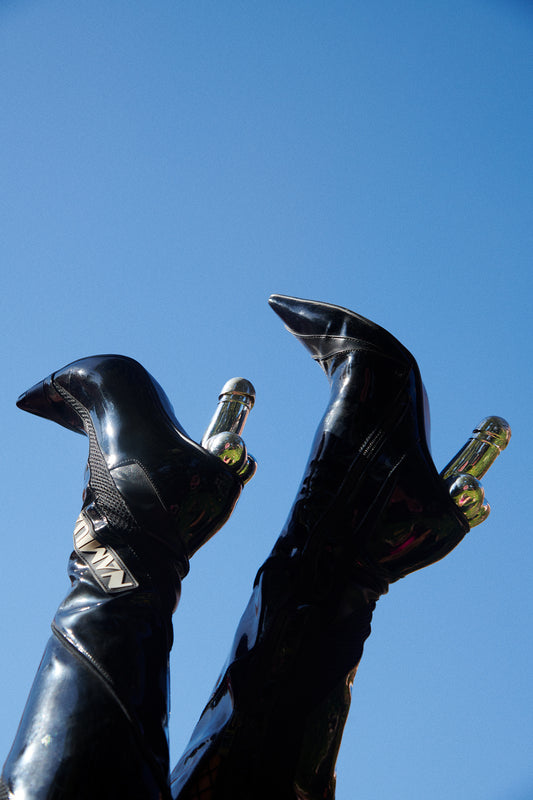 A photograph of a pair of black shiny boots with silver penis heels set against a blue sky, titled 'Penis Pumps'.