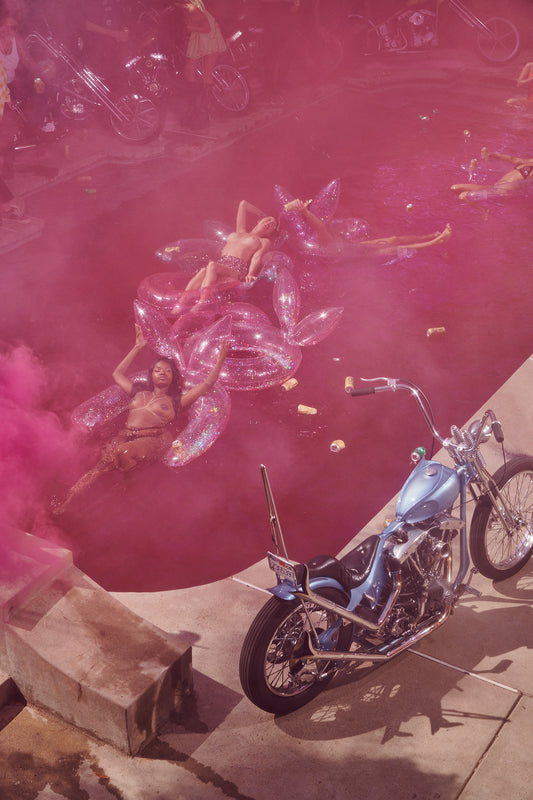 The image displays an art print titled "Pink Haze," capturing the essence of a countercultural pool party from the 1960s or 1970s. The artwork features a Radcliffe & Coopers motorcycle, notable for its chopper style, parked near a pool, with a background filled with pink smoke or mist, suggesting a vibrant, perhaps even psychedelic atmosphere.
