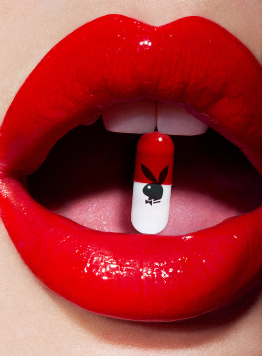 Close-up of red lips with a pill bearing the Playboy logo placed between them.