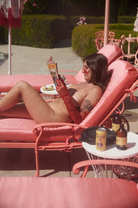 An art print depicting a person in a hangover reading a magazine while lying on a pink lounge chair, with coors beer s on a side table. The setting is a poolside area with pink furniture.