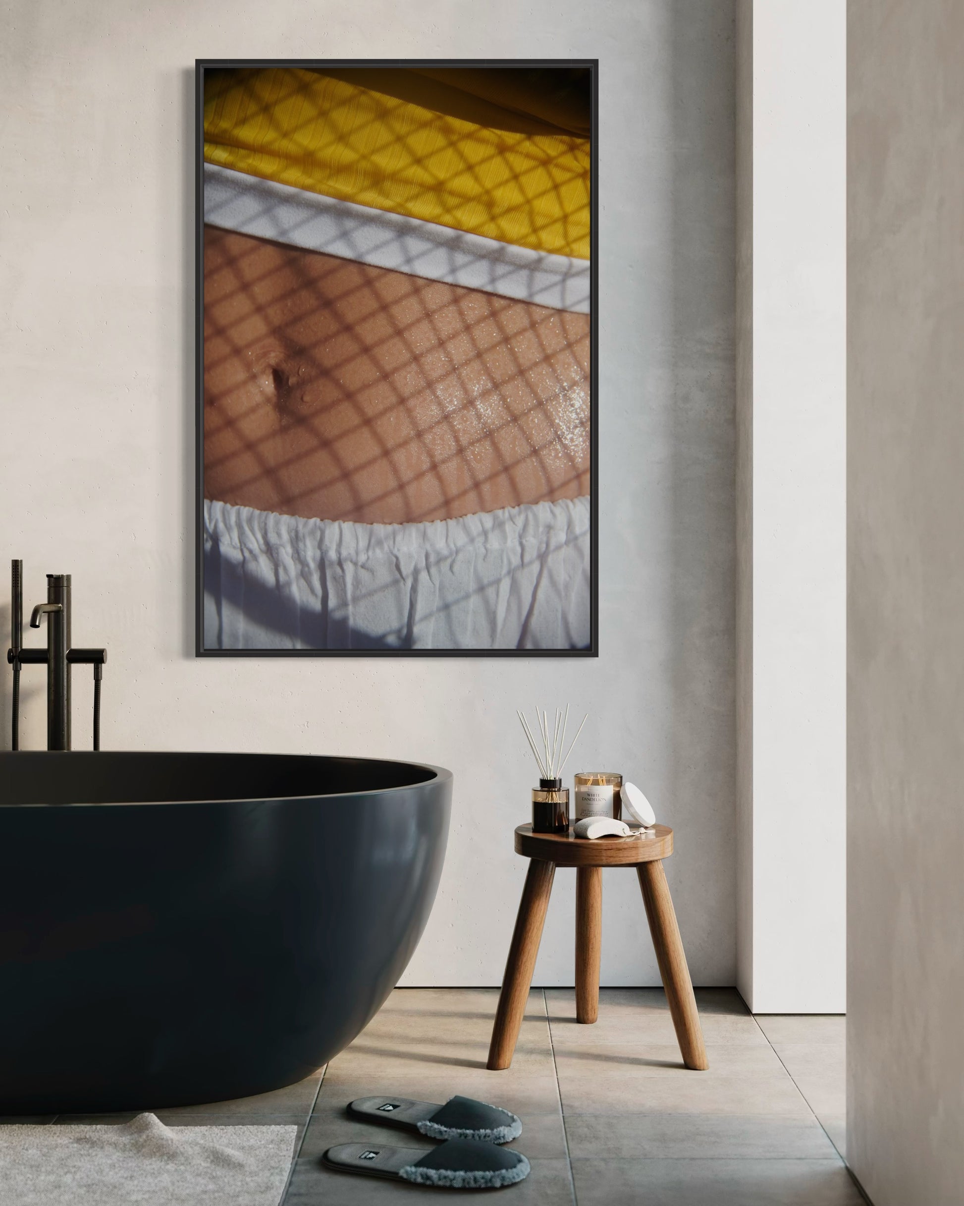 A modern bathroom with a black freestanding bathtub, wooden stool, and a framed photograph of a woman's stomach with sweat beads on it and the shadows of a tennis racket projected onto it.