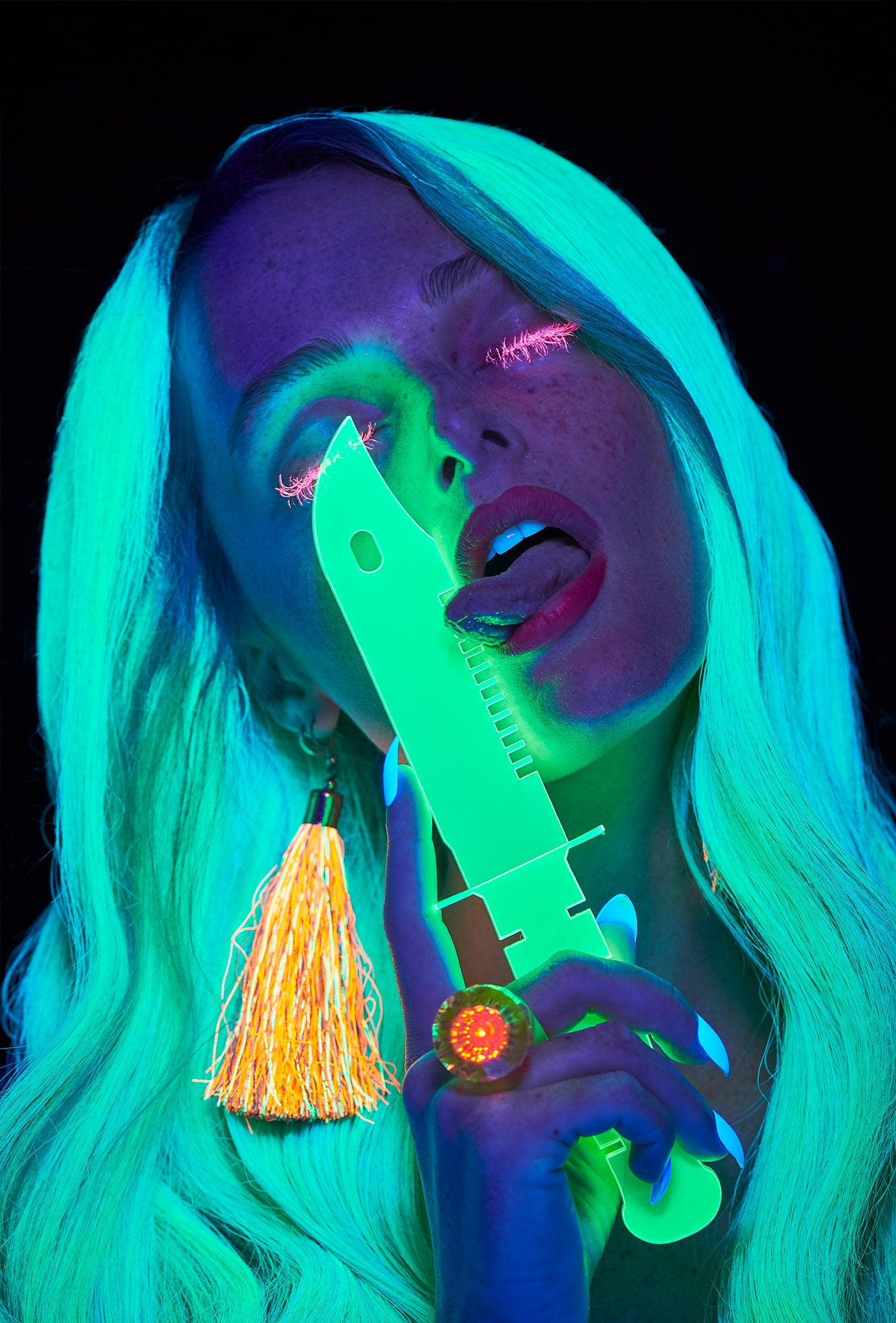 Artistic image of raver party club girl with pink mascara eyelashes and neon green hair licking a glow-in-the-dark knife