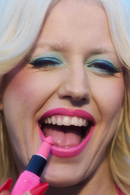 A portait of Amy Tayor (Amyl and the Sniffers) wearing blue eyeshadow and applying pink lipstick while winking and smiling.