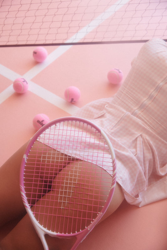 Woman lying down on a pink tennis court wearing a pink tennis mini dress surrounded by pink tennis balls with a tennis racket on her exposed buttocks.