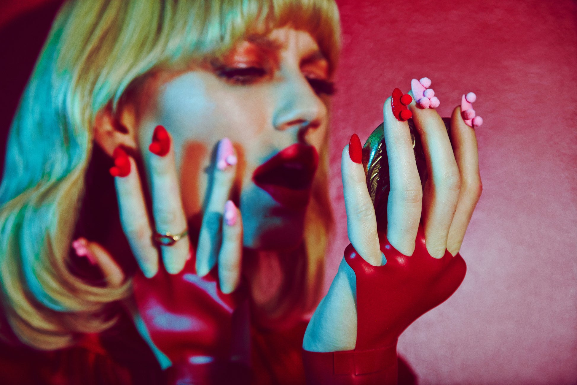A close-up of a woman's hands with a red manicure and pom-pom details, holding a camera up to her face, with a vibrant red and pink color tone photographed by top beauty photographer located in Los Angeles, Jamie Nelson.