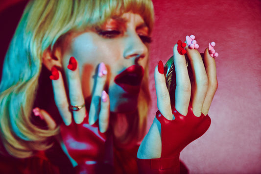 A close-up of a woman's hands with a red manicure and pom-pom details, holding a camera up to her face, with a vibrant red and pink color tone photographed by top beauty photographer located in Los Angeles, Jamie Nelson.