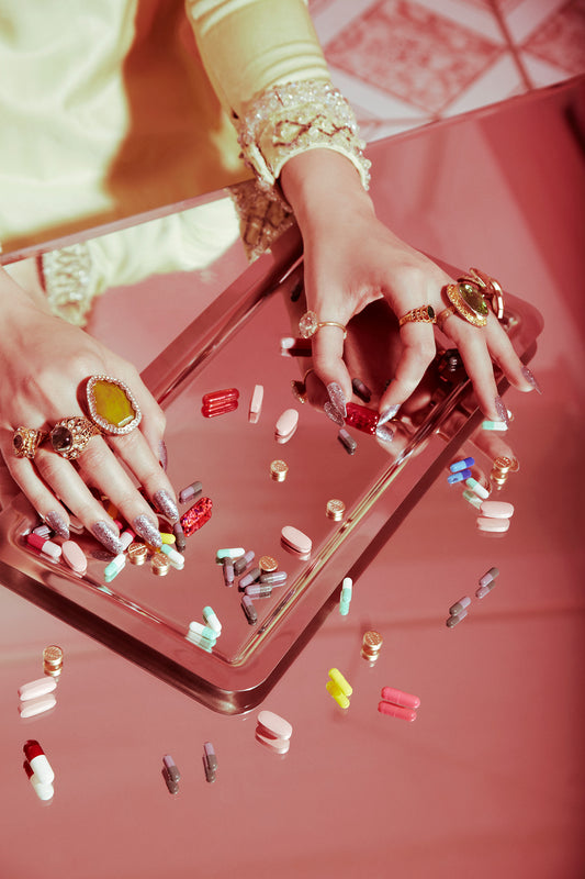A person with a glitter manicure counting colorful pills on a reflective surface, with a vintage aesthetic.