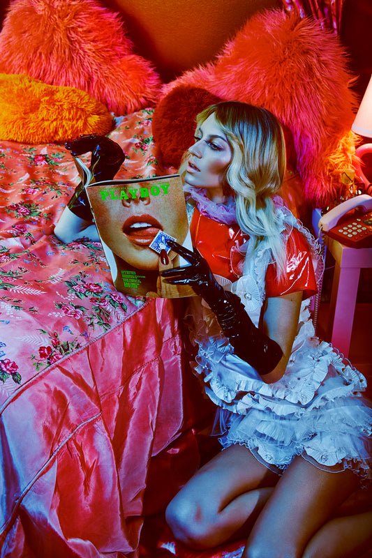 A stylized fine art photograph depicting a woman in a coquette aesthetic, seated in a vintage bedroom with pink and orange tones, reading a vintage Playboy magazine.