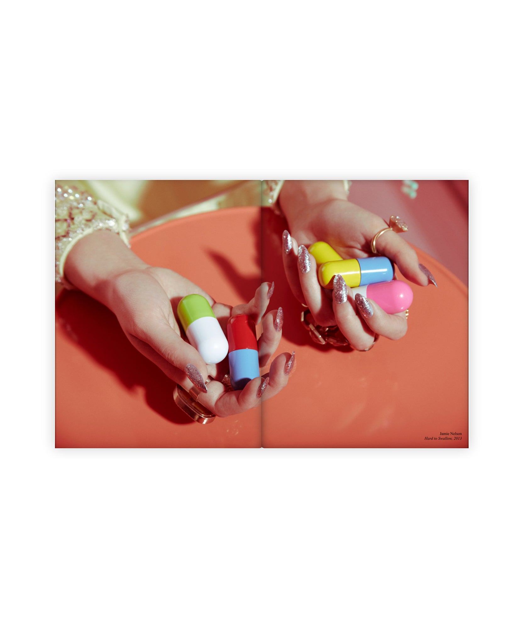 An artistic vintage style photography print in an art book by Jamie Nelson. The woman wears large rings and glitter nail polish as she holds colorful giant pills in her hand against a coral surface.